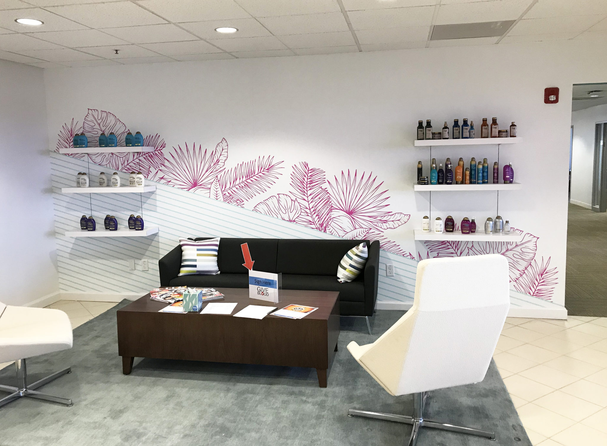 White seating area with white chairs, gray couch, slate gray rug, white shelves with colorful bottles of hair products on them on an accent wall with a decorative pattern of aqua-colored stripes and pink leaves and palm fronds Vogue International offices