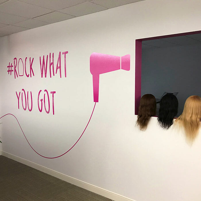 White wall with bright pink vinyl cling graphic of a hairdryer with the words Rock What You Got next to it. Hairdryer graphic is pointed at a windowsill with three wig-wearing mannequin heads in it.